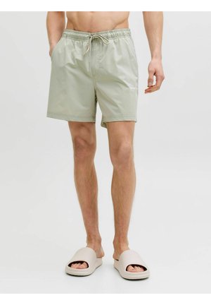 Mężczyzna w jasnozielonych szortach ze ściągaczem i beżowych sandałach na płaskiej podeszwie stoi na jednolitym białym tle.