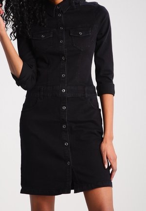 Nainen, joka pukeutunut mustaan napitettuun farkkumekkoon, jossa on kaksi rintataskua ja käännetyt hihat, seisoo yksivärisen taustan edessä.