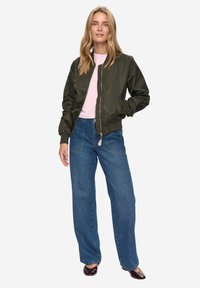 Bomber bunda olivově zelené barvy s žebrovanými manžetami, ve stylu spojená se světlou růžovou košilí a modrými širokými džíny. Model stojí s rukama v kapsách.