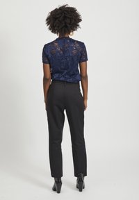 Mörkblå spetsblus med blommönster, korta ärmar och knäppning bak, kombinerad med svarta skräddarsydda byxor och klackskor.