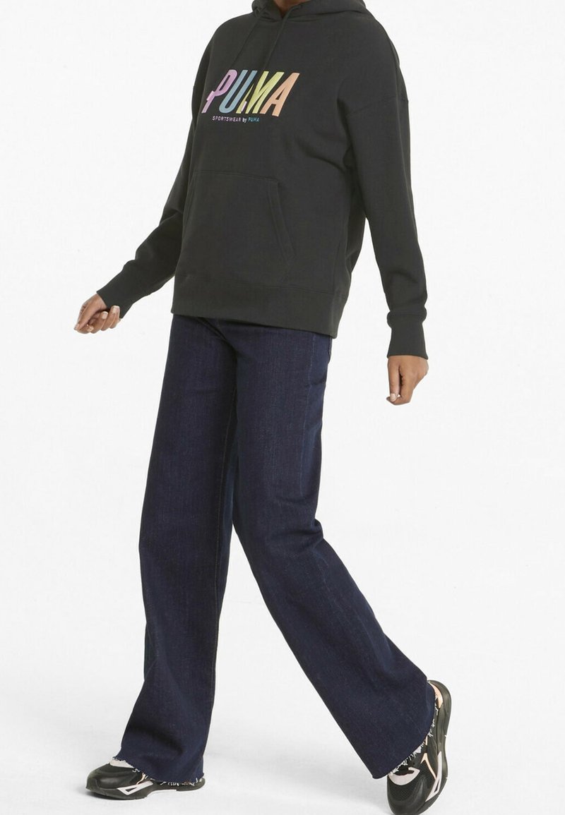 Sweat à capuche noir avec un logo "PUMA" multicolore, poche avant ; associé à un jean large bleu foncé et des baskets noires.