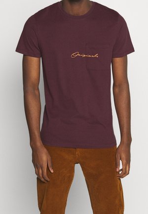 Mężczyzna noszący ciemnofioletowy t-shirt z krótkim rękawem i kieszonką na piersi z pomarańczowym napisem "Originals," w połączeniu z brązowymi sztruksowymi spodniami.