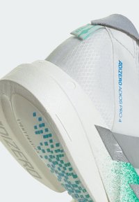 Talons de chaussure de course blanche et grise avec des éclaboussures turquoise, texte bleu « ADIZERO ADIOS PRO 4 » et semelle extérieure texturée avec motif de grip bleu.