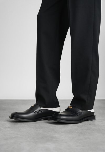 Mocassins pretos de couro com um acabamento suave, apresentando uma tira e um detalhe decorativo, combinados com calças pretas sob medida.