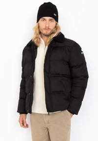 Sort pufferjakke med krave-design og lynlås, der har en glat overflade og et diskret logo på ærmet. Båret over en cremet sweater.