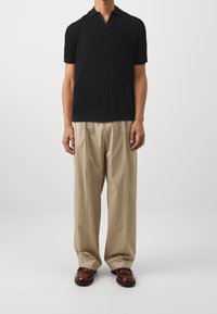 Mustat lyhythihaiset neulepolo, jossa on teksturoitu kuvio, yhdistettynä kermaisiin pleatti-housuihin ja ruskeisiin nahkaläppäkenkiin.