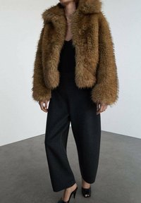 Hnedý kožúšok z umelé kožušiny s kapucňou, skrátený strih a voľné čierne nohavice na mieru. Nosí sa s čiernymi sandálmi na opätku s otvorenou špičkou na šedivej podlahe.