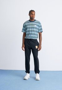 Mann, der auf einem blauen Boden steht, ein gestreiftes blau-grünes T-Shirt, schwarze Jogginghosen mit einem Patch und weiße Sneaker trägt und zur Seite blickt.