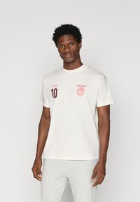 Κρεμ χρώματος βαμβακερό T-shirt με κοντά μανίκια, με μπορντώ κείμενο "O Rei" και "10" στο αριστερό στήθος, και ένα λογότυπο επ badge από πάνω.