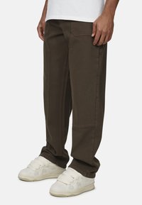 Brązowe bawełniane spodnie cargo o luźnym kroju, z bocznymi i tylnymi kieszeniami oraz przeszyciami. Noszone z jasno-beżowymi sneakersami.