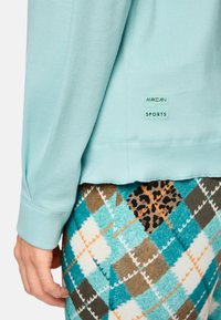 Light blue ribbed long-sleeve top with MARC CAIN SPORTS tag, paired with patterned leggings featuring turquoise and brown diamond shapes and leopard print.