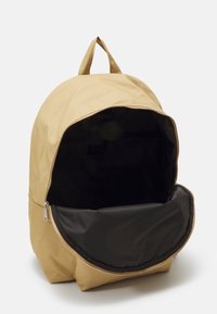 Beige rugzak met een ronde vorm, een ritsluiting aan de buitenkant en een gestructureerd oppervlak. De binnenkant heeft een zwarte voering en een ruime compartment.