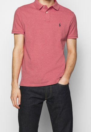 Mann trägt ein kurzärmeliges pinkes Poloshirt mit kleinem dunklem Logo auf der linken Brust, in dunkelblaue Jeans gesteckt, eine Hand in der Tasche.