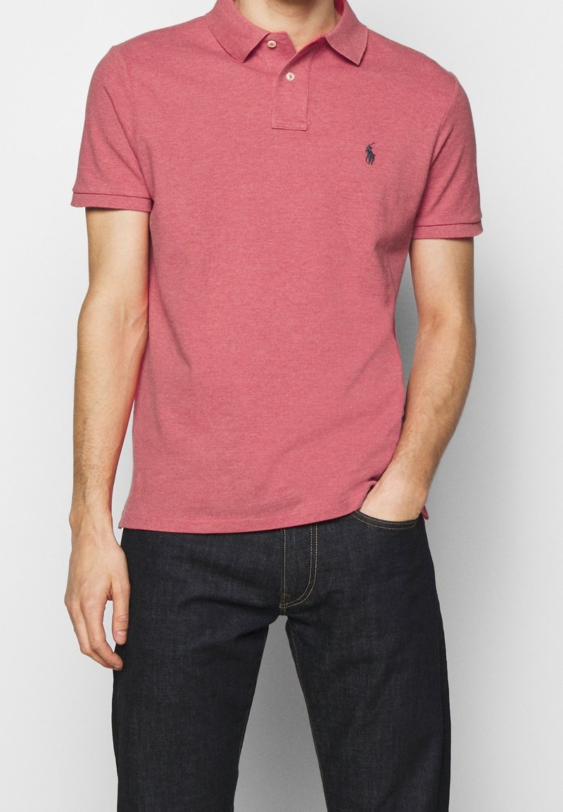 Homme portant un polo rose à manches courtes avec un petit logo sombre sur le côté gauche de la poitrine, rentré dans un jean bleu foncé avec une main dans la poche.