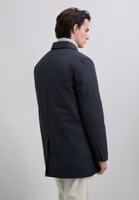 Mężczyzna w ciemnoniebieskim płaszczu na jasnoszarym golfie, odwrócony plecami, na jednolitym jasnoszarym tle.