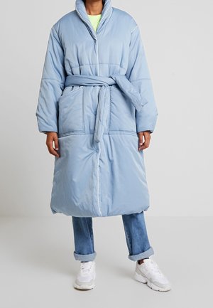 Cappotto lungo imbottito di colore azzurro chiaro con vita svasata, ampie tasche laterali e colletto alto. Bordature bianche che accentuano le cuciture.