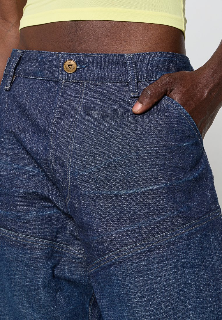 Jeans w ciemnoniebieskiego denimu o teksturowanym wykończeniu, z przednią kieszenią i metalowym zapięciem. Widoczna nitka wzdłuż szwów.