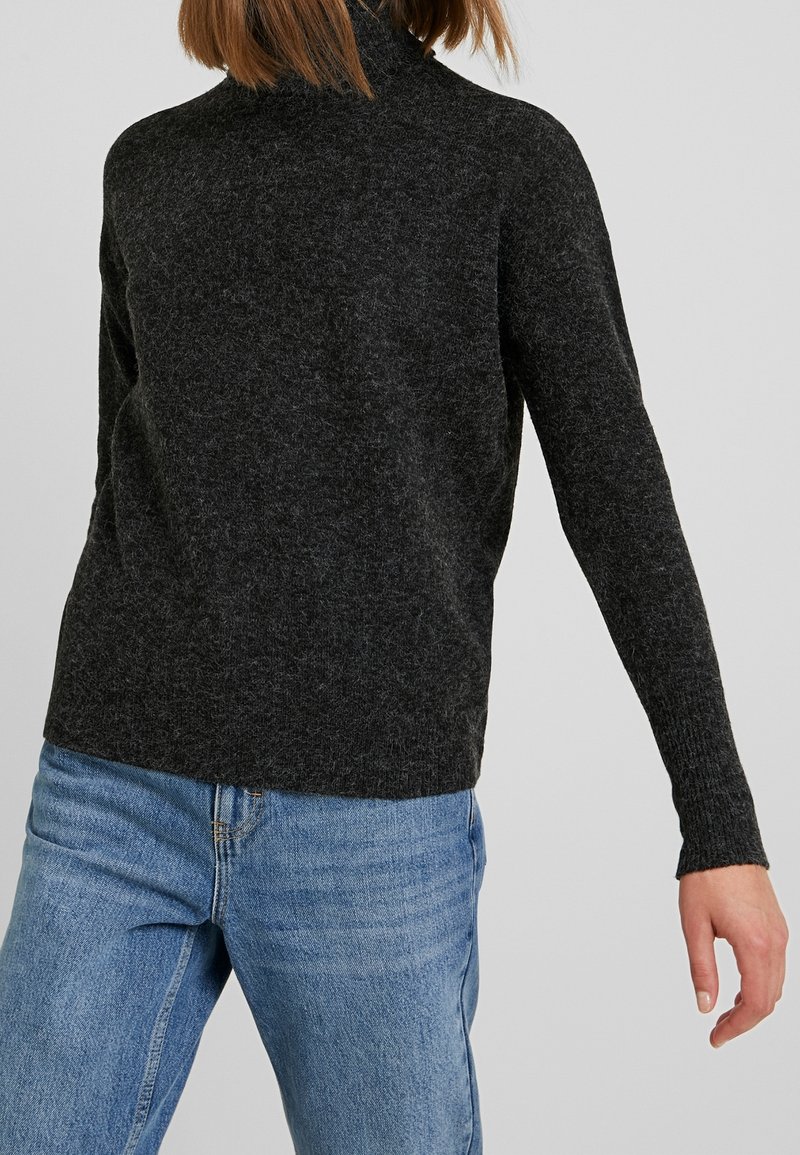 Suéter de cuello alto en gris oscuro, de tejido suave, con mangas largas, dobladillo redondeado y un corte holgado, combinado con jeans de un azul claro.