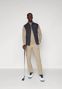 Golfoutfit bestaande uit een marineblauwe mouwloze jas, een beige overhemd met lange mouwen, beige broek en witte golfschoenen met blauwe accenten. Model houdt een golfclub vast.
