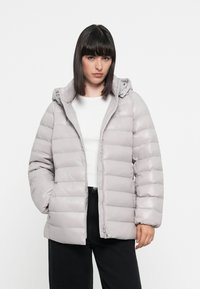 Šedá pufrová bunda s kapucňou, prešívaný dizajn, predný zips a elastické manžety. Nosená cez biele tričko, spárovaná s čiernymi nohavicami.