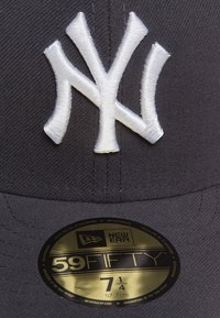 Gorra de béisbol negra con un logo NY bordado en blanco. Cuenta con una visera plana y una etiqueta dorada que indica la talla 7 1/4. Está fabricada con un tejido texturizado.