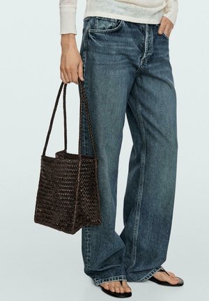 Personne portant un jean bleu ample et un haut blanc à manches longues, portant un sac fourre-tout tissé foncé et des tongs noires.