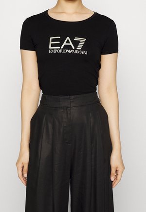 Vrouw draagt een zwart T-shirt met korte mouwen met zilveren EA7 Emporio Armani-logo en zwarte, hoog getailleerde, wijde broekspijpen.
