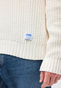 Kremfarget strikket genser med en teksturert vaffelmønster. Har ribbestrikkede mansjetter og kant, med en blå og hvit merkeetikett nederst.