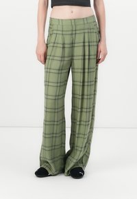 Person wearing loose-fitting olive green plaid pants and black Puma slides, standing against a plain white background.