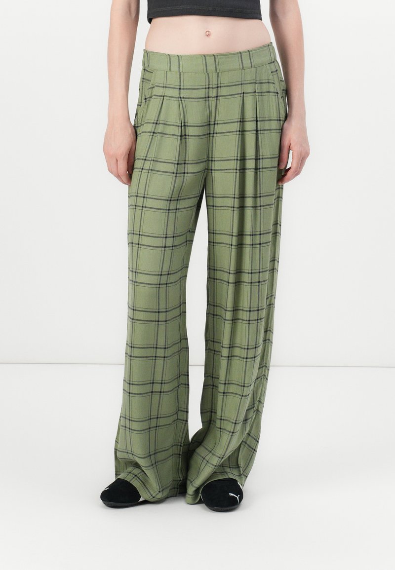 Person wearing loose-fitting olive green plaid pants and black Puma slides, standing against a plain white background.