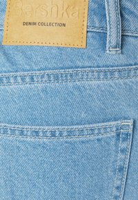 Ljust blå denimtyg med en synlig bakficka med en läderlapp präglad med "Bershka Denim Collection." Stygn detaljer längs kanterna.
