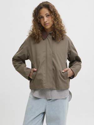 Veste marron en tissu vert olive, avec deux poches latérales et une coupe décontractée, superposée à une chemise à carreaux et un jean clair.