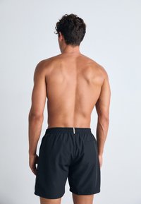 Black swim trunks with an elastic waistband and a gold accent. The fabric appears smooth with a relaxed fit, shown from a rear view.