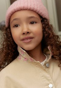 Chaqueta beige con un cuello de estampado floral, que presenta herrajes plateados. La modelo lleva un sombrero de punto rosa claro, mostrando su cabello rizado.