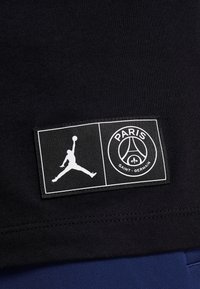 Etiqueta de tela negra con la silueta blanca de Jumpman y el logo del Paris Saint-Germain que incluye la Torre Eiffel y símbolos de la flor de lis.