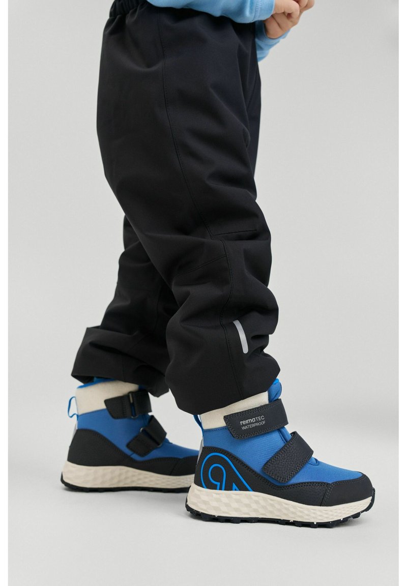 Niebieskie wodoodporne buty z czarnymi akcentami, paskami na rzepy, zaokrąglonym noskiem i teksturowaną podeszwą. Połączone z czarnymi, wodoodpornymi spodniami.