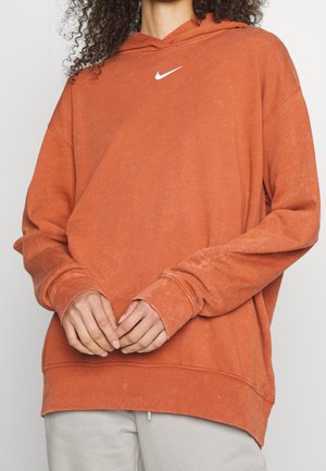 Person wearing oversized burnt orange hoodie with white Nike logo, hands loosely clasped in front, paired with light-colored pants.