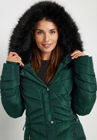 Giacca puffer verde con cappuccio in pelliccia eco-friendly nera, caratterizzata da una texture trapuntata, zip frontale e chiusure a scatto, progettata per calore e stile.