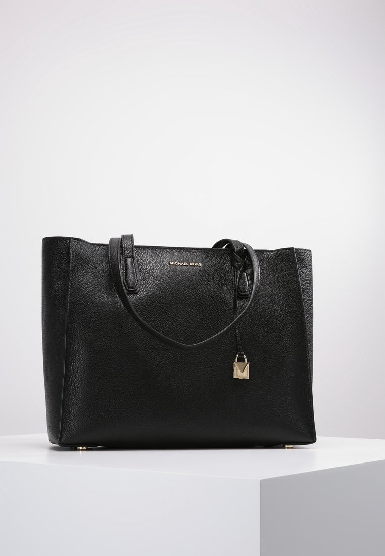 Sac cabas en cuir noir Michael Kors avec doubles poignées et un petit charm en forme de cadenas doré, exposé sur un socle blanc.