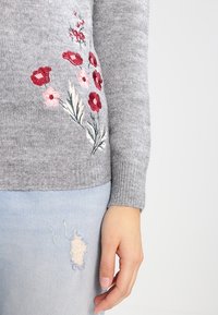 Main portant un jean bleu clair déchiré et un pull gris avec une broderie florale rouge et rose sur la manche et le côté.