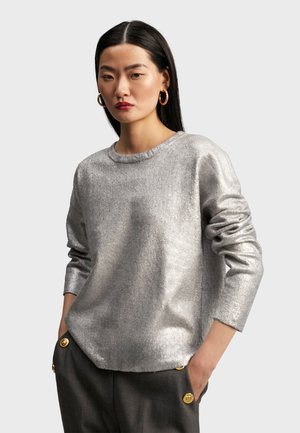 Silver metallic long-sleeve top with a round neckline and slightly frayed edges. Soft texture with a relaxed fit, paired with tailored grey trousers.