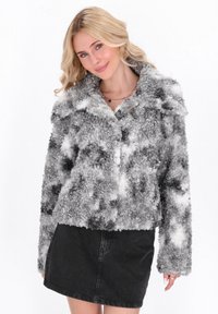 Joven mujer con cabello rubio vestida con una chaqueta de forro polar texturizada en gris y blanco y una falda de mezclilla negra, sonriendo ligeramente.