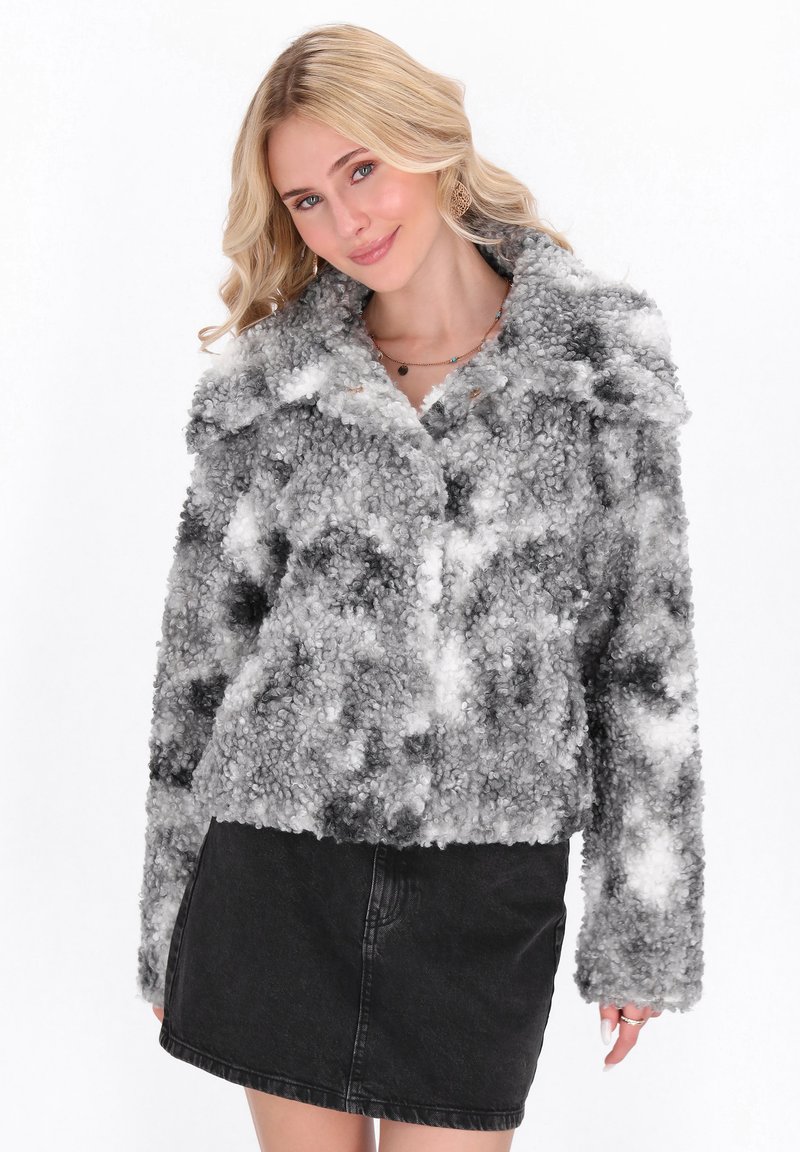 Joven mujer con cabello rubio vestida con una chaqueta de forro polar texturizada en gris y blanco y una falda de mezclilla negra, sonriendo ligeramente.