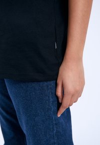 Camiseta de algodón negra con una etiqueta lateral, combinada con pantalones de mezclilla azules. Enfoque en la textura de la tela y el contraste de colores. La mano descansa sobre la pierna del pantalón.