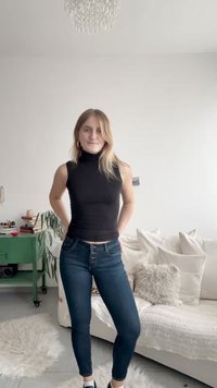 Giovane donna con dolcevita nero senza maniche e jeans scuri in piedi in un soggiorno luminoso con divano bianco e tavolino verde.