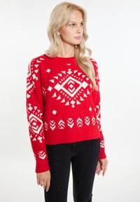 Pulover tricotat roșu, cu un model geometric alb și o textură moale și pufoasă. Guler rotund și mâneci lungi. Asortat cu pantaloni negri.