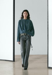 Volně střižená halenka v barvě modrozelené s detaily na zavazování, doplněná šedými zvonovými džínami. Materiál působí měkce a má viditelnou texturu. Černá obuv.