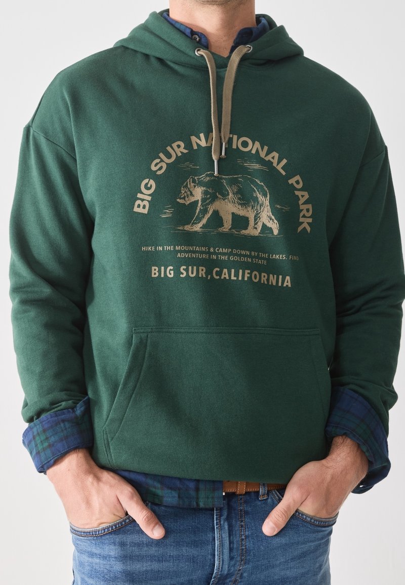 Sudadera verde bosque con el gráfico "Parque Nacional Big Sur" en beige, con una ilustración de un oso, capucha con cordones y bolsillo frontal.