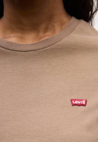 Chemise à col rond marron avec un patch logo Levi's rouge sur la poitrine supérieure, portée par une personne au teint brun moyen.