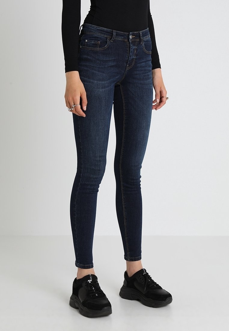 Person som bär mörkblå skinny jeans, svart långärmad tröja och svarta klumpiga sneakers, stående mot en enkel vit bakgrund.